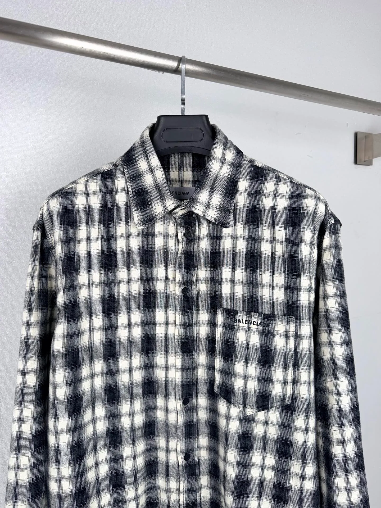 BALENCIAGA/balenciaga Black White plaid pocket embroidered logo shirt jacket The technical difficulties of this plaid shirt focus on fabric finishing, plaid matching and component sewing details. The fabric has been sanded and distressed to create a fine velvet feel on the surface, changing the original luster of the fabric and making the Black and White plaid colors transition softly. At the same time, the drape properties of the fabric itself are retained, taking into account the looseness of the upper body and the silhouette of the body, without appearing soft and shapeless. The biggest test of plaid garments comes from the alignment of the pieces. The front and rear body, large and small sleeves, and chest pockets are all independent pieces. In the cutting stage, the horizontal and vertical axes of the plaid need to be aligned, and the external chest bag is cut separately. The plaid on the bag surface needs to form a visual coordination with the main body of the garment. Cutting and typesetting will produce additional fabric loss, and there are high requirements for the cutting accuracy of the cutting bed typesetting and workers. The collar adopts a double-layer combined structure, and the collar surface and collar seat are double-stitched with even and regular stitching spacing to ensure the lapel's long-lasting shape and resistance to warping and deformation after repeated bending. The placket is sewn with regular topstitching and paired with Black matte buttons. The button points need to be aligned with the checkered lines to avoid damaging the overall visual order. The splicing of the cuffs also takes into account the grain direction, and the overlocking process reinforces the seams to improve the durability of the garment. After the entire garment is sewn, it also undergoes a garment washing process to further strengthen the Retro texture of the fabric, unify the tone and texture of the entire garment, weaken the harshness of industrial production, and finally present a relaxed Retro garment effect. Size: 46 48 50 52